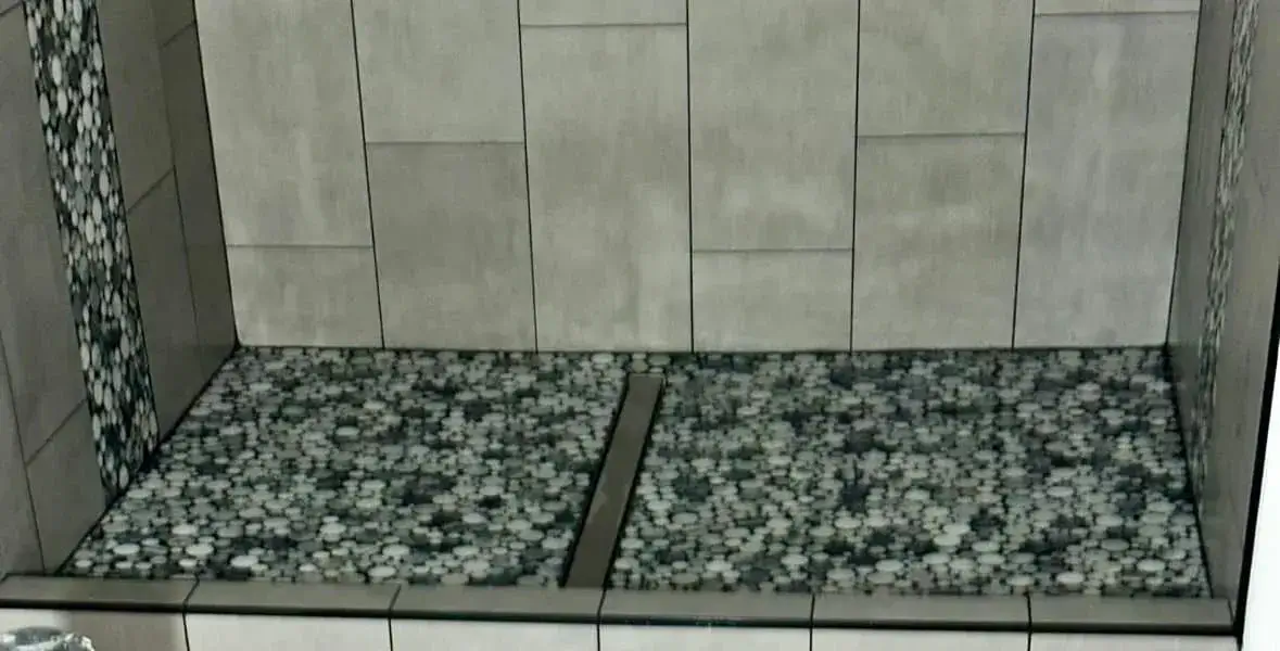 Detail of custom pebble-mosaic shower floor with linear drain and large-format gray porcelain wall tile by Zona Floors