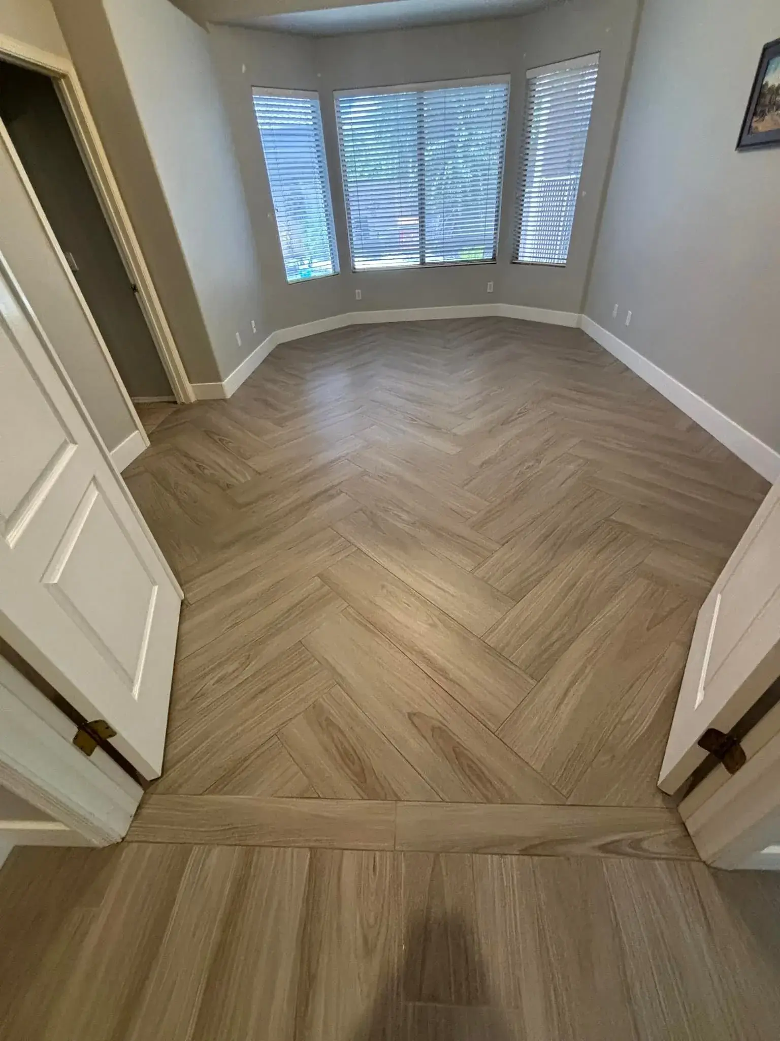 Light oak herringbone wood-look flooring installed in an Arizona bay-window sitting nook by Zona Floors, transitioning to straight-plank flooring in the adjoining hallway