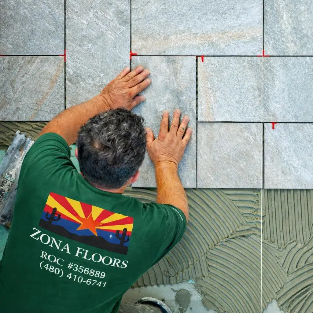 Top-down view of a licensed Zona Floors installer (ROC #356889) in branded shirt placing large-format stone tile with red leveling spacers in an Arizona home